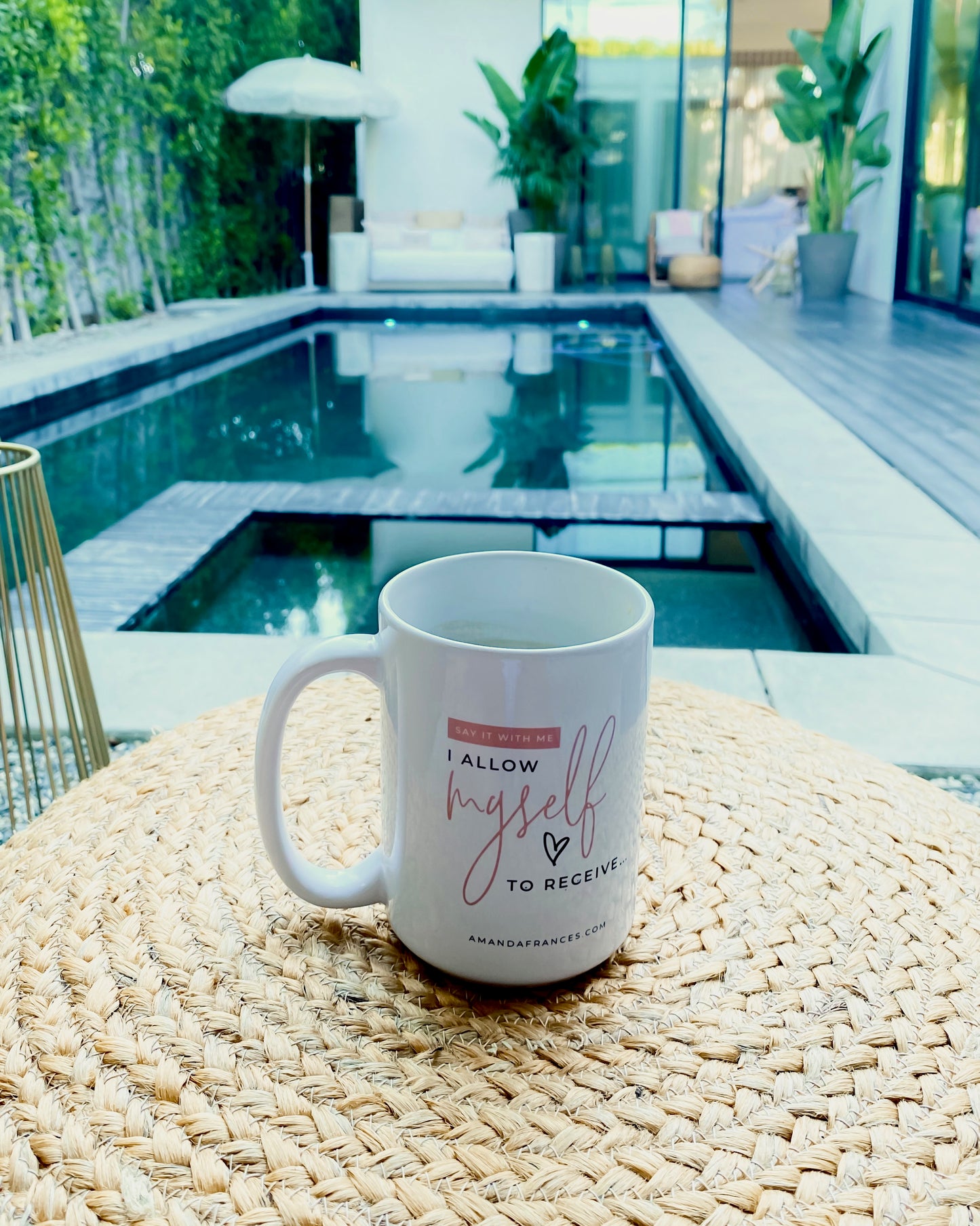 White glossy mug with the text: 'Say it with me, I allow myself to receive' and 'amandafrances.com' sitting on a table .y the pool